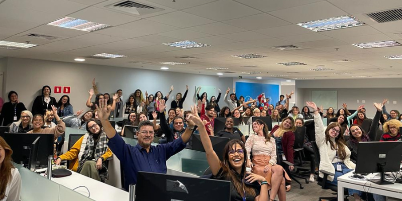 Group photo of enthusiastic Airswift employees in an office setting, smiling and waving during a team event.