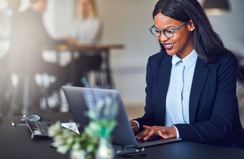 Smiling young businesswoman working on a laptop at her desk in a bright modern office with colleagues in the background