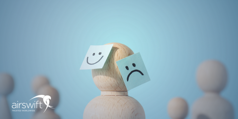 Wooden figure with two sticky notes on its head, one with a sad face and one with a happy face, against a blue background with other blurry wooden figures.