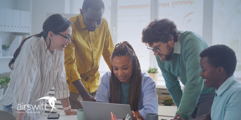 Five colleagues of different ethnic backgrounds gathered around a laptop, discussing training needs analysis data. The team is smiling and engaged, with one person typing on the laptop while others lean in, offering suggestions.