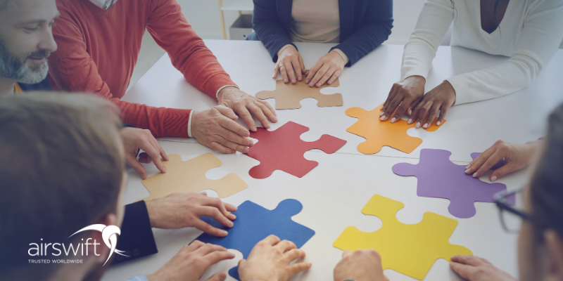 A diverse group of colleagues gathered around a table, each placing their hands on colourful puzzle pieces. The image symbolizes teamwork and collaboration. 