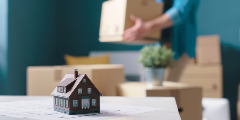 A miniature of a house on top of a table. In the back, someone is moving out, carrying boxes.